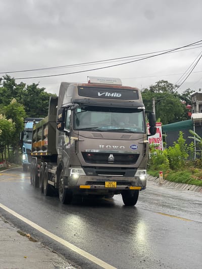 The image captures a rainy day scene on a paved road in Sơn Dương Commune, Vietnam. A large, brown dump truck is prominently featured in the foreground, driving on the right side of the road. The truck has the "HOWO" logo visible on its grille, and a license plate displaying "90C 103.06". Another blue truck is visible behind the brown one, also driving on the right. The road is wet, with reflections of headlights and the sky on its surface. Trees line the sides of the road, and buildings are visible in the background. The sky is overcast and grey, indicating a cloudy or stormy day. Electrical wires crisscross above the road. A red and white marker post is visible on the right side of the road. There are no visible people in the image.