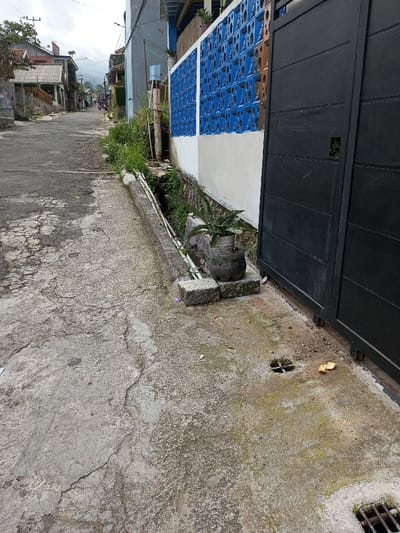 The image depicts a residential street scene in Viengthong, Laos, under an overcast sky. The foreground on the right features a black, ribbed metal sliding gate. Adjacent to the gate, a dark, cylindrical planter containing a green leafy plant rests on two stacked square concrete blocks on the cracked concrete pavement. The pavement exhibits areas of green moss or algae growth and contains a small, square open drain and a discarded yellow object. A metal-grated drain is visible at the bottom right. A narrow concrete curb runs parallel to the street on the left, bordering a vegetated drainage ditch containing white pipes. Behind this ditch, a wall consists of a white lower section topped with blue-painted, openwork concrete breeze blocks. A utility pole and a structure with a water tank are visible behind the wall. The main street, paved with cracked asphalt or concrete, recedes uphill, flanked by various residential buildings of diverse construction. Overhead utility lines are present. Distant hills or mountains are visible in the background. No discernible individuals are present in the immediate scene.