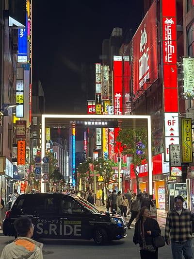 The image captures a vibrant street scene at night in Shinjuku, Japan. Neon signs dominate the scene, advertising businesses such as Taito Station, restaurants, and shops. Pedestrians fill the sidewalks, engaged in various activities. A black car with "S.RIDE" prominently displayed on its side is parked along the street. The overall atmosphere is that of a bustling entertainment and commercial district in a major city.