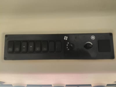 The image shows a close-up, eye-level shot of a control panel mounted on the ceiling of a vehicle, likely a tractor or construction equipment. The panel is black and rectangular, with several buttons and two rotary knobs. From left to right, there are six rectangular buttons. The first button has a symbol resembling a light bulb. The second button has a symbol that appears to be "90". The third button has a triangle with a line through it. The fourth button has a similar symbol to the third. The fifth button has a windshield wiper symbol. The sixth button is blank. To the right of the buttons, there is a fan symbol with an arrow, and a rotary knob with settings numbered 1, 2, and 3. Next to that is a snowflake symbol, followed by another rotary knob. Finally, on the far right, there is a square blank button. The control panel is attached to a light-colored, textured surface, possibly the interior headliner of the vehicle. The image does not depict any specific location in the city of Krylov, Russia, as it is a close-up of an indoor control panel.