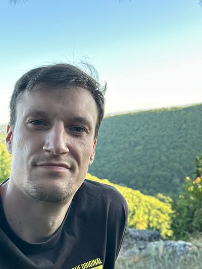 A man with short, dark hair and stubble is looking at the camera. He is wearing a dark t-shirt. He is outdoors, with a blurred background of green trees and a bright sky. The image appears to be a selfie, taken from a low angle. The man's face is in sharp focus, while the background is out of focus.