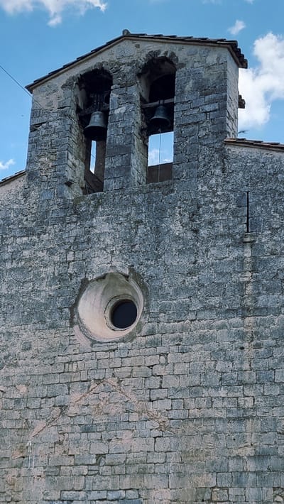 The image shows the facade of a stone building with a bell tower. The bell tower has two arched openings, each containing a bell. Below the bell tower is a circular window with a dark interior. The stone blocks of the building are weathered and uneven. The sky above is blue with some scattered clouds. The building is located in Montagut i Oix, Spain.