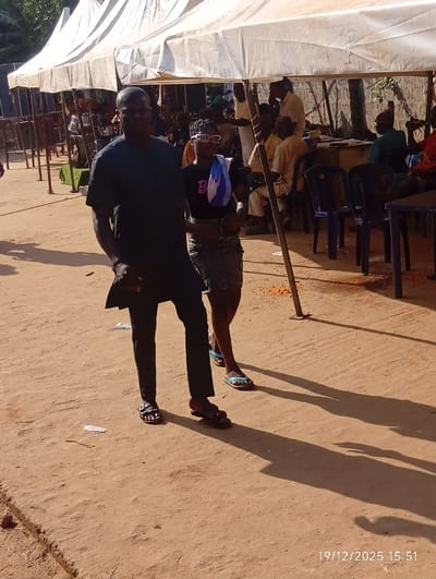 An outdoor scene in Isinyi-Nando, Nigeria, depicts a gathering under white canopy tents on a clear, sunny day. The foreground features two individuals walking on reddish-brown dirt ground. On the left, a dark-skinned male wears a dark short-sleeved top, dark trousers, and dark sandals. To his right, a dark-skinned female wears eyeglasses, a black short-sleeved shirt with a "Barbie" logo, a dark patterned skirt or shorts, and light blue flip-flops, with a blue and white striped fabric draped over her left shoulder. Long shadows are cast to the right on the uneven ground. In the background, numerous individuals, predominantly men in light-colored attire, are seated on blue plastic chairs or standing under several white tents. Dark tables are visible among the seated figures. The environment is characterized by dry, unpaved ground with scattered light-colored debris and a visible timestamp of 19/12/2025 15:51 in the lower right corner.