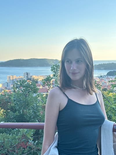 A young woman stands on a balcony overlooking the city of Čučuci, Montenegro. She has shoulder-length brown hair and is wearing a dark green camisole and a thin necklace. She is holding a light-colored blanket over her arm. The background shows a coastal city with buildings and a body of water, with hills in the distance under a clear blue sky. Lush green foliage is visible in the foreground and around the balcony. The lighting suggests it is late afternoon or early evening.