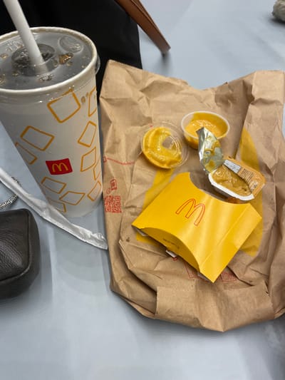 A close-up, elevated view depicts fast food items from McDonald's resting on a light grey, smooth surface, consistent with a table, within an airport in Israel. On the left, a white paper cup containing a dark beverage with ice and a white straw features the red McDonald's logo and a pattern of white and orange-yellow rectangles. Condensation is visible on the cup's exterior. Adjacent to the cup, a crumpled white paper straw wrapper is present. To the right, an open, crumpled brown paper bag, displaying a QR code and "mcdonalds.com," contains a yellow McDonald's branded cardboard carton. Three distinct containers of yellowish sauce are visible within or near the bag: one clear plastic container with its lid partially displaced, one small open white plastic cup, and one opened foil packet. A dark grey or black small bag or purse with a metallic zipper pull is partially visible on the far left of the frame. No individuals are depicted.