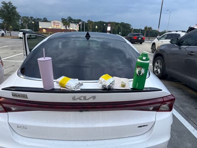 The image shows the rear of a white Kia K5 car in a parking lot. Two foil-wrapped food items and a small container are placed on napkins on the car's trunk lid between a purple tumbler on the left and a green water bottle with a Boston Celtics logo on the right. In the background is a building with the words "Micky Finn's" on it. The sky is overcast. There are other cars parked nearby. The location is Florence, South Carolina, United States.