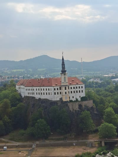An aerial view of the Děčín Castle, located in Děčín, Czechia, shows the fortress perched on a rocky outcrop. The castle features a long white building with a red-tiled roof and a prominent black tower with a clock. Surrounding the castle is dense green foliage and a distant mountainous landscape under a cloudy sky. Below the castle, a pathway with a bridge crosses a body of water, and there are a few people visible walking or cycling. Two portable toilets are also present in the foreground. The scene captures the historical architecture of the castle amidst a natural and urban environment.