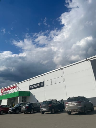 A low-angle, sideways shot captures a building facade with a parking lot in front, under a dramatic sky. The building appears to be a retail establishment, possibly a supermarket, with large white panels and a green awning over an entrance. A sign with red Cyrillic text is visible, which reads "Пятёрочка" (Pyaterochka), a Russian supermarket chain. Another sign above the entrance displays "ПИВОВАРНЯ" (Pivovarnya), meaning "Brewery." Several cars are parked in a row on the asphalt in front of the building. One car, a silver Kia SUV, has a license plate ending in "HY 18." The sky is a mix of deep blue and dark, stormy grey clouds, suggesting a dynamic weather system, possibly late afternoon or early evening given the lighting. The overall scene conveys a sense of everyday commerce under a striking natural backdrop.