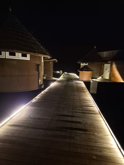 Captured at night in Bodufolhudhoo, Maldives, the image shows a dark, angular view of architecture. A section of a multi-story structure with brown paneling and white-framed windows is visible, juxtaposed against a long, illuminated wooden wall. The illumination is provided by a strip of light running along the top edge of the wall. The sky is completely black, creating a high-contrast scene. There are no people, animals, or signs of activity present. The architectural style appears modern and functional. The orientation of the image is rotated clockwise, making it appear as if the buildings are leaning into each other.