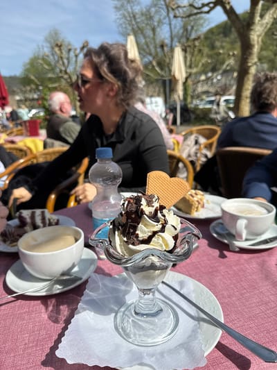 The scene depicts an outdoor café setting, likely in Cochem, Germany, given the architecture and style. The primary focus is a dessert sundae served in a stemmed glass, topped with whipped cream, chocolate syrup, and chocolate shavings, adorned with a heart-shaped waffle. The dessert sits on a white plate lined with a delicate paper doily. A long silver spoon rests on the plate. To the right of the sundae is a white cup of coffee with a saucer and another spoon. Beside it, there's a slice of cake.
A woman with sunglasses is visible in the background, seated at the table. Beside her is a water bottle. Other patrons are seated nearby, their faces partially visible. Wicker chairs are used in the café. The table is covered with a pink tablecloth. The background includes trees and possibly buildings, indicating the café's location in an urban or semi-urban environment. The time of day is likely daytime, suggested by the daylight and shadows.