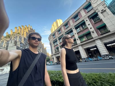 This full-shot image, taken from a low-angle perspective, captures two individuals standing on a city street, likely in the Putuo District of China, under a bright, clear blue sky.

In the foreground, a woman is positioned on the left side of the frame, wearing a black crop top and black shorts. She has sunglasses perched on her head and appears to be looking upwards towards the camera. To her right, a man wears a dark, sleeveless top and black sunglasses, with a black bag strap diagonally crossing his chest. He also looks towards the camera.

Behind the individuals, a city street is visible with a double yellow line. A row of shared bicycles, typically seen in Chinese urban settings, is parked along the curb. Beyond the street, lush green bushes and trees line the pavement.

The background features two prominent buildings. On the left, a multi-story building displays a facade of red brick sections interspersed with white or beige trim, featuring multiple windows, some adorned with small green planters. The word "COLMO" is clearly visible on its ground-level facade. Further in the distance and to the right, another taller building with a striking golden or yellowish exterior and decorative architectural elements on its roofline stands against the clear sky, showcasing a modern urban environment. The bright lighting and lack of shadows suggest a sunny day, possibly late morning or afternoon.