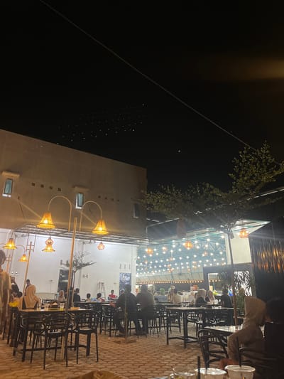 The image depicts a night scene at an outdoor dining establishment in Matangjareung, Indonesia. The setting is illuminated by numerous hanging lights, creating a warm and inviting atmosphere. A building with a light-colored facade and several windows is visible in the background. The foreground features rows of tables and chairs, with patrons seated and conversing. Several people are visible, some of whom are wearing headscarves. The ground is paved with a textured material, possibly cobblestones. A tree with lush foliage is positioned to the right of the frame. The overall impression is that of a lively social gathering in a well-lit, open-air restaurant.