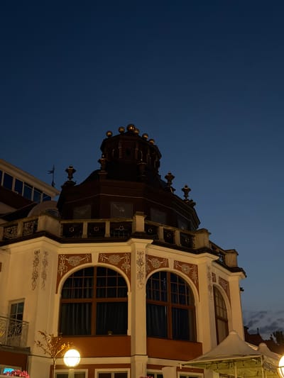 The image captures an ornate building in Sopot, Poland, under a twilight sky. The building features a prominent dark dome with decorative gold accents and several smaller decorative finials. The facade is painted white with intricate reddish-brown detailing around arched windows, some of which are covered by dark curtains. A decorative balustrade runs along the upper levels. In the foreground, a streetlamp with a circular globe illuminates the area. The overall scene suggests a charming, possibly historic, urban environment at dusk.