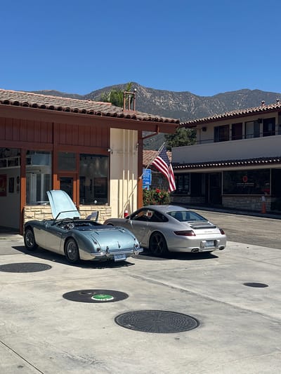 The image displays a sunlit outdoor scene in Montecito, United States, featuring two parked automobiles in front of buildings with Spanish Colonial Revival architecture, set against a backdrop of distant mountains.

In the foreground, a light blue classic Austin-Healey 3000 convertible is parked, oriented away from the viewer. Its hood is open and propped up. The soft top is down, revealing a dark interior. To its right, a silver Porsche 911 Carrera coupe (likely 997 generation) is parked, also oriented away from the viewer, featuring dark multi-spoke wheels.

The vehicles rest on a light gray concrete pavement embedded with several circular manhole covers, one centrally displaying a green insert.

Behind the cars, a single-story building with a brown wooden facade, large glass windows, and a terracotta tile roof is visible on the left. An American flag hangs vertically from a pole attached to this building's right corner, above a blue sign. A security camera is mounted on the building's facade. To the right, a two-story building with cream-colored stucco walls and a similar terracotta tile roof is present. Its ground floor windows partially reveal an interior sign with "ARCHITECTS" visible.

The background consists of a clear blue sky and distant, dry, vegetated mountains. Strong shadows on the ground indicate a bright, midday setting. No individuals are present in the scene.