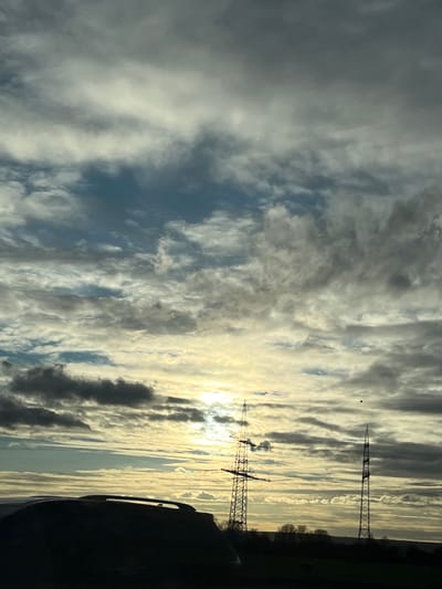 This image depicts an outdoor landscape observed from within a vehicle, situated in the vicinity of Bergheim, Germany. The scene is dominated by a cloudy sky with patches of blue. The sun, partially obscured by clouds, is visible as a bright glare in the lower left-center, backlighting the foreground and midground elements.

In the immediate foreground, the dark silhouette of the roof and upper rear section of a vehicle, possibly an SUV or minivan with roof rails, occupies the bottom left quadrant.

In the midground, two distinct towers are prominent. To the left, a large lattice-steel power transmission tower (pylon) with multiple cross-arms and visible power lines stands silhouetted against the bright sky. Further to the right, a second, taller, slender lattice tower with a pointed top is also silhouetted. A distant treeline and rolling terrain are visible along the horizon between and beyond the towers.

In the upper right quadrant of the sky, above and slightly to the left of the second tower, a small, distinct black circular object is present.

No individuals, specific actions, or direct interactions are visible within the frame. The overall lighting conditions suggest either early morning or late afternoon.