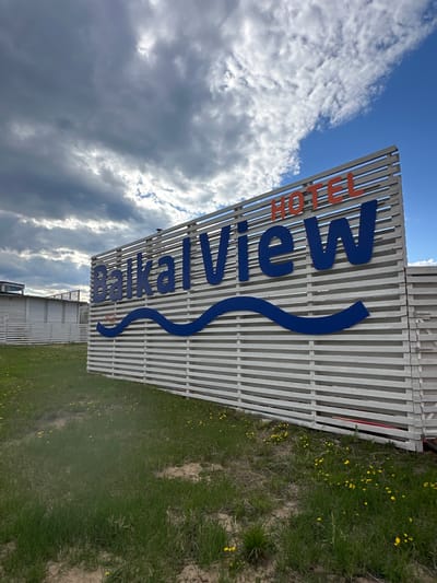 A ground-level, tilted, and full-frame photograph depicts a sign for "Baikalview HOTEL" in Khuzhir, Russia. The sign consists of white horizontal slats, with the word "HOTEL" in orange, vertical block letters. Below "HOTEL," the word "Baikalview" is written in large, blue, vertical block letters, with a wavy blue line underneath. The sky, with cumulus clouds, occupies the upper left portion of the image. Ground cover, with patches of grass and small yellow flowers, is visible on the right. A white building with horizontal slats is partially visible below the sign.