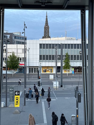 Captured from an elevated perspective, the image portrays a street scene in Ulm, Germany. The Ulm Minster's spire rises above the urban landscape. A modern building with a grid-like facade stands prominently, with businesses visible on the ground floor. Pedestrians cross the street at a crosswalk. Street furniture such as lamp posts and signage is in view, including a yellow "TAXI" sign with a downward arrow. The street is paved and marked with lane dividers. The perspective is framed by architectural elements. The weather appears to be clear with a blue sky and light clouds.