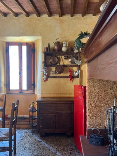 This image shows the interior of a room, likely a kitchen or dining area, in a building located in Sinalunga, Italy. The room features a window with wooden shutters, partially open, allowing natural light to enter. The walls are painted in a textured yellowish-orange hue, decorated with simple floral patterns. Displayed on the wall are a collection of copper pots, pans, and kitchen utensils arranged on a wooden rack. Other decorative items include dried peppers and greenery. A wooden cabinet with drawers and doors is visible. A red structure extends from the top of the cabinet. A chair and a table partially occupy the lower portion of the frame. The ceiling exhibits exposed wooden beams. A fireplace is placed in the upper right portion of the scene, along with metal fire tools. The floor has a mosaic tile pattern.
