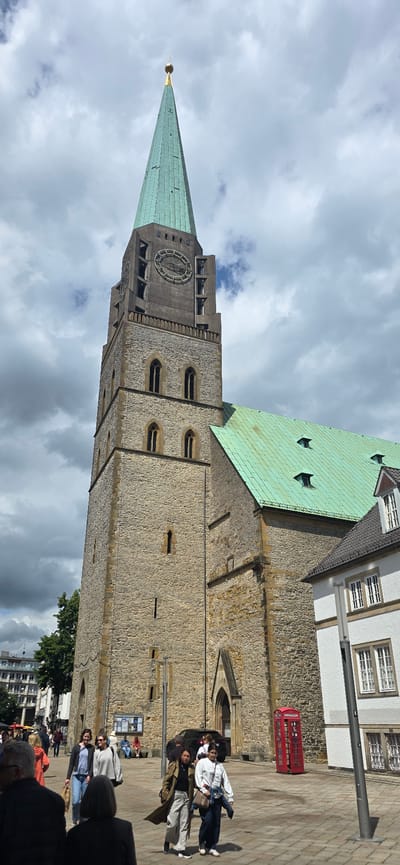 This is a ground-level, vertical, full-body shot of the historic Old Town Hall (Altes Rathaus) in Bielefeld, Germany. The building features a tall, stone tower with a distinct green, peaked roof topped with a gold spire. The tower has a large, round clock face and several arched windows. Adjacent to the tower is another section of the building with a green, sloped roof that has dormer windows. In the foreground, several people are walking on a paved square in front of the building. A classic red British-style telephone booth stands near the building's entrance. The sky is partly cloudy.