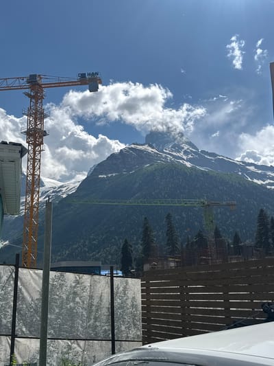 This image captures a construction site nestled against a majestic mountain backdrop. The scene is dominated by towering mountains, their peaks capped with snow and their slopes covered in dense green forest. The sky is a vibrant blue, dotted with fluffy white clouds, suggesting a clear and pleasant day.

In the foreground, construction cranes are prominently visible. An orange crane, labeled with the text "LIEBHERR," stands tall, its lattice structure reaching towards the sky. To its left, a green crane is also at work. The construction site itself appears to be in progress, with scaffolding and building materials evident in the background, partially obscured by a wooden fence and a white tarp. The presence of construction activity suggests a development project is underway in this scenic mountain region.

The image evokes a sense of progress and ambition, with human endeavors taking shape against the enduring power of nature. The time of day appears to be daytime, likely late morning or early afternoon, given the bright sunlight casting shadows and illuminating the scene. The location context of Dombay, Russia, places this construction within a region known for its natural beauty and outdoor recreational opportunities.