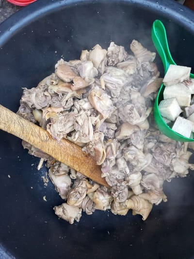 A high-angle shot captures a black pot filled with cooked chicken and diced white yams, along with a wooden spoon and green ladle. Steam rises from the cooked chicken pieces. This image appears to be taken in Isunba, Nigeria.