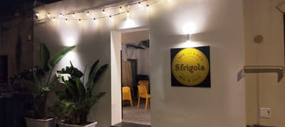 An outdoor scene at night in San Vito Lo Capo, Italy, features a building with a white stucco exterior, illuminated by a string of globe lights overhead and two wall-mounted globe lights. To the left, large green plants in white planters are visible, along with dark trash bins. An open doorway leads into a dimly lit interior where yellow chairs and decorative screens are present. On the exterior wall, a black square sign with a yellow circular logo is mounted, featuring Italian text that reads "L'ARANCINA A VISTA Sfrigola MADE IN SICILY". A metal pipe and a small electrical box are also attached to the wall.