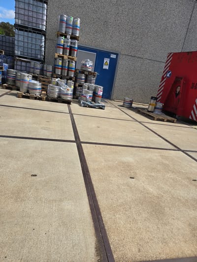 In the city of Melle, Belgium, a concrete yard features several stacks of metal kegs and large industrial containers. In the background, a grey speckled building stands tall, with a blue door and a yellow warning sign. To the right, a large red container is visible, marked with white stripes and a drawing of a person. The yard is sectioned by dark, embedded metal strips in the concrete. The overall scene depicts an industrial or storage area under a bright sky with scattered clouds.