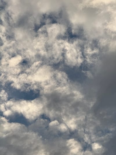 The image is a close-up, high-angle shot of a cloudy sky. The clouds are varied in density and color, ranging from bright white where illuminated by the sun to dark gray in shadowed areas. There are patches of blue sky visible between the clouds, suggesting it is daytime. The clouds appear to be cumulus or stratocumulus, with a textured, somewhat turbulent appearance. The lighting suggests the sun is positioned somewhere above and to the right of the frame, casting shadows and highlighting the edges of the cloud formations. There is no discernible text, people, or objects in the frame, making the sky itself the sole subject. The setting is outdoors, specifically looking upwards at the sky. The overall impression is one of natural atmospheric conditions, potentially indicating changeable weather. The location context provided is Дзержинец-83 Снт, Russia, but there are no specific visual cues within the image to confirm this location. The time of day appears to be daytime, and the weather is cloudy with glimpses of blue sky.