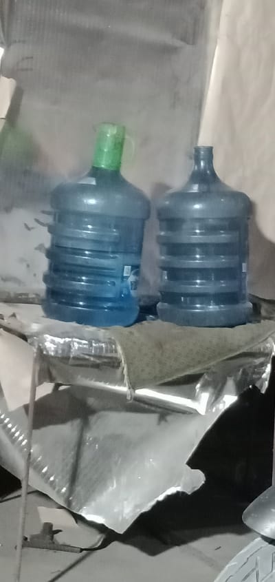 Two large, blue plastic water jugs are positioned on a surface that appears to be a shelf or table. The jug on the left has a green cap and a visible label. The jug on the right has a dark cap. The background is indistinct and appears to be a wall with some textured or uneven surface. Below the jugs, there is reflective material, possibly aluminum foil, and a dark fabric or tarp. The lighting is dim, casting shadows. The scene is indoors, likely in a storage area or similar setting. The image does not contain any information that would allow for identification of a specific location in Tanjungan, Indonesia.
