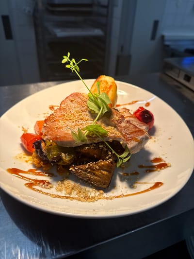 A close-up, overhead shot showcases a white plate with a gourmet dish. The dish features pieces of sliced meat, possibly pork or veal, topped with finely chopped nuts and herbs. Alongside the meat are roasted vegetables, including eggplant and tomatoes, and a golden-brown pastry or bread component. A glistening caramel-colored sauce is artfully drizzled across the plate, adding a touch of elegance. The lighting suggests a professional kitchen environment, possibly during food preparation or service. The background is blurred, hinting at stainless steel equipment and a generally dark, indoor setting. There is no visible text in the image.