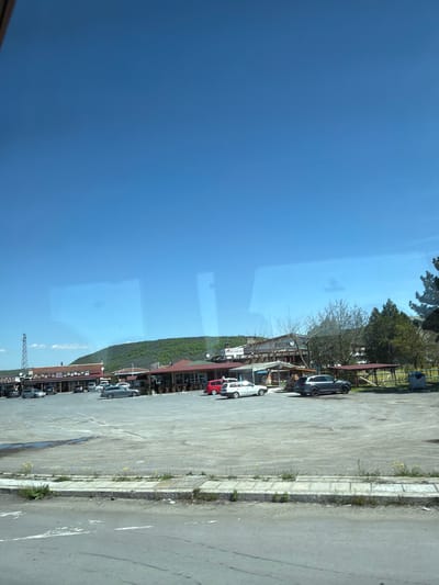 The image shows a street view, possibly taken from inside a vehicle, in Gorno Aleksandrovo, Bulgaria. The sky is clear and blue. A row of buildings lines one side of the street, with cars parked along the roadside. The buildings appear to be commercial establishments, possibly restaurants or shops, judging from the signage and awnings. The road is paved and appears to be moderately used. Vegetation, including trees and shrubs, is visible near the buildings and along the roadside. The scene seems to capture a regular day in the city, with no specific interactions or actions prominently visible. The environment is typical of a small town or village.