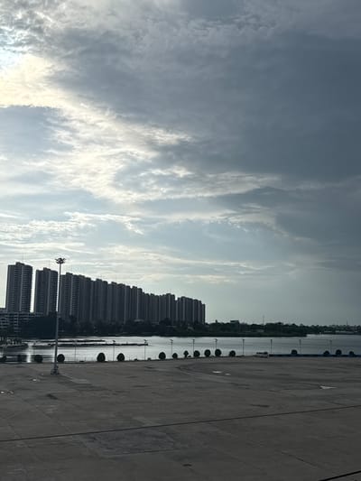 This image depicts an outdoor urban scene. In the foreground is a large, paved area with distinct grid lines, likely a plaza or parking lot, with scattered circular planters and lampposts. Beyond this area, a body of water is visible, with a shoreline lined with more lampposts and a row of dense, tall buildings in the mid-ground. The buildings appear to be residential or commercial high-rises. The sky is overcast with dramatic, textured clouds, and a bright patch of sunlight breaks through on the left side. The scene is devoid of people. Based on its visual characteristics and common urban development, it is plausible this location is within Pak Kret City Municipality, Thailand, characterized by its waterfront developments and modern high-rise architecture.