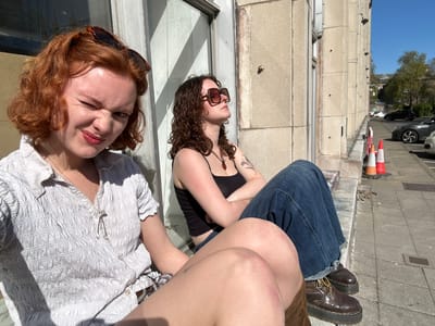 The image shows two young women outdoors in a sunny urban setting, consistent with the context of Dundee, United Kingdom. They appear to be sitting or leaning on a concrete ledge or wall, with a building and a street in the background.
The woman on the right has red/auburn hair and a fair complexion. She is wearing a light-colored, possibly white or off-white, textured button-up shirt with smocked detailing. A silver chain necklace is visible around her neck. Her sunglasses are pushed up onto her head. She looks directly at the camera with a playful expression, one eye slightly squinted or winking, and has red lipstick.
The woman on the left has dark, curly hair. She is wearing a black spaghetti-strap top, dark blue denim jeans with a distressed or frayed hem, and dark brown lace-up boots with thick soles. She wears dark, square-framed sunglasses on her face and has visible tattoos on her arms and shoulders. She is looking to her left, away from the camera.
The background features a multi-story, light-colored building with multiple rectangular windows. In front of the building, a street is visible with parked cars, including a silver hatchback and a black vehicle. Two traffic cones, one red and white and another orange and white, are standing on the pavement. Distant trees and a clear blue sky are visible in the upper left background.
The strong sunlight casts distinct shadows, indicating a bright and clear day. The women seem to be casually relaxing and posing for a photograph. There is no visible text in the image. The scene suggests a casual outing or break in a city environment.
