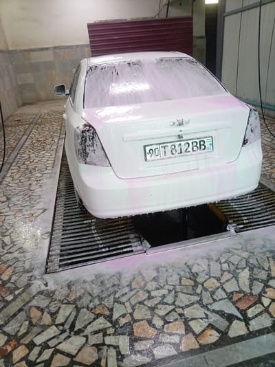 The image shows the rear of a white Chevrolet car covered in white and pink foam at a car wash in Bagat, Uzbekistan. The car is positioned over a metal grate drain system. The floor is tiled with irregularly shaped stones in shades of gray, beige, and brown. Soapy water is visible on the floor and the drain. The car's license plate, reading "90 T 812 BB UZ", is clearly visible. The background suggests an indoor car wash facility with tiled walls and a corrugated metal roof. There are no people visible in the image.