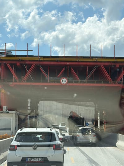 The image captures a view from within a vehicle driving under a red metal construction structure, possibly a bridge or overpass, on a sunny day with a blue sky and white clouds. A circular sign indicating a height restriction of 4.5 meters is visible on the underside of the structure. In the foreground, the rear of a white Citroën C3 car with the license plate XPT-1740 is prominent. Behind it, other vehicles, including a silver car and a truck with a crane, are visible in traffic. The scene appears to be a roadway in the Polichni Municipal Unit of Greece, characterized by the construction and the surrounding infrastructure.
