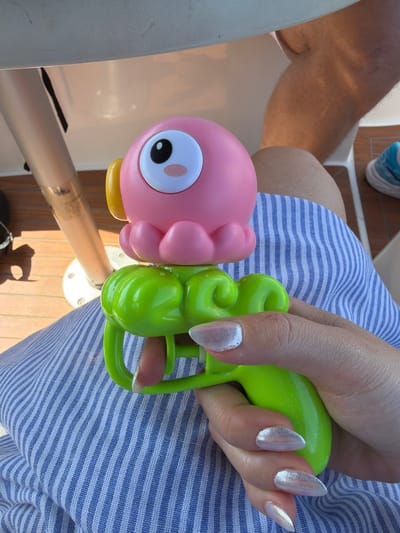 The image shows a close-up of a person's hand holding a bright green toy water gun. The water gun is shaped like a cartoon character with coiled green appendages. A pink, cartoonish octopus toy with a yellow beak and a large eye is attached to the front of the water gun. The person's fingernails are long and painted with a shimmery, iridescent polish. They are wearing a blue and white striped garment. In the background, a person's leg, clad in blue and white sneakers, is visible, resting on a wooden deck. The setting appears to be outdoors, possibly on a boat or a patio, with warm lighting suggesting it is daytime. The overall impression is a playful and relaxed scene. No text is clearly legible on the water gun.