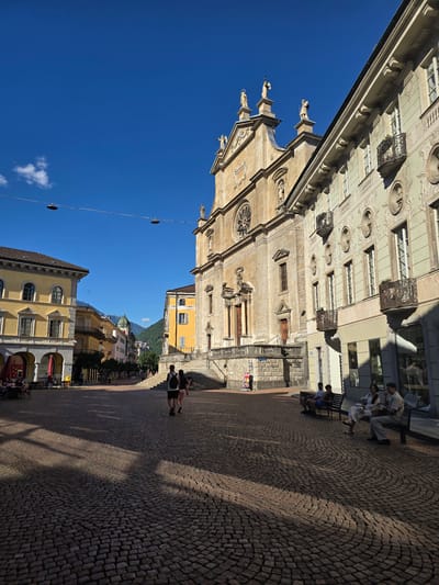 The image depicts a sunny day in an outdoor plaza, likely in Bellinzona, Switzerland. The scene is dominated by historical architecture, including a grand, light-colored stone building with intricate carvings and decorative balconies. Adjacent to it, another building features a yellow facade. In the distance, more colorful buildings and a lush green hillside with a domed structure are visible, suggesting a picturesque European town.
Several people are present in the plaza. Some are seated on benches, while others are walking along the cobblestone ground. The lighting suggests it is daytime, with distinct shadows cast by the buildings and people, indicating a bright, clear sky. The cobblestone paving is a prominent feature, adding to the historic atmosphere. The overall mood is relaxed and tranquil, with individuals enjoying the pleasant weather and surroundings. There is no visible text in the image.