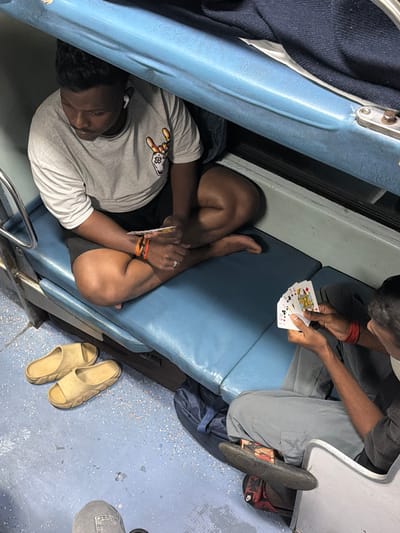 In Valsad, India, two individuals are seated on blue padded benches, likely within a train compartment. The person on the left, wearing a light gray t-shirt and black shorts, sits cross-legged, holding a deck of playing cards in their right hand and a ring on their left ring finger. They have earbuds in their ears. The person on the right, facing away from the camera and wearing a gray sweatshirt and pants, holds several playing cards in their hands. Both individuals appear to be engaged in a game of cards. A pair of beige slip-on sandals rests on the speckled blue floor to the left of the frame. A dark blue bag is visible behind the individual on the right. The environment suggests a confined space with tiered seating, characteristic of train travel.