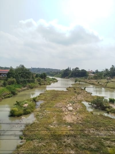 The image shows a view of a river landscape in Đại Ninh, Vietnam. The river is wide and murky, dividing into multiple channels with grassy islands. The sky is overcast. In the background, buildings and trees line the horizon. Wires are visible in the foreground.
