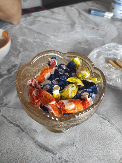 A clear, intricately patterned glass bowl is filled with an assortment of individually wrapped candies. The candies are primarily in orange, blue, and yellow wrappers. Some of the orange wrappers have text that appears to read "ORANGE" and "BAVANSULI." The yellow wrappers display images of lemons and some have text that might read "LEMON." The blue wrappers appear to have text including "PINK" and "Coconut." The bowl is placed on a textured, light-colored surface that resembles a marble tabletop with prominent grey veining. In the background, a white ceramic bowl with an orange rim is partially visible on the left. To the right of the glass bowl, a clear plastic bag contains some elongated, light-brown items, possibly biscuits or crackers, and a blurred blue and white object is visible further in the background. A person's arm and shoulder are faintly visible in the upper left corner of the frame. The scene is lit by natural light, casting soft shadows. The location is identified as Некрасовка, Kazakhstan.