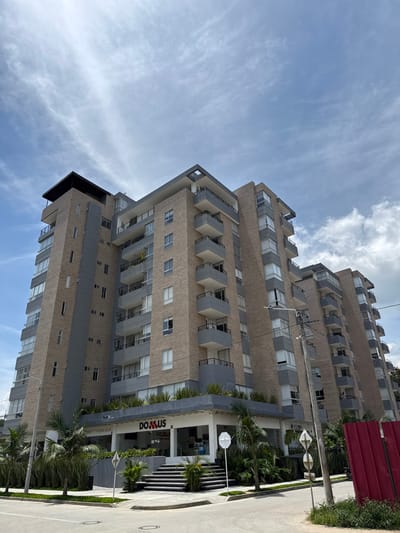 This image depicts the "DOMUS Apartamentos" building, located at Calle 3 Sur #2-13 in Pitalito, Huila, Colombia. The multi-story building, approximately nine to ten floors tall, features a modern architectural design with light brown brick facades and dark grey paneling. Each floor includes multiple balconies with dark railings and grey concrete bases. The ground floor houses commercial establishments, prominently displaying a "DOMUS" sign in red lettering. A tiered entrance with steps leads up to the commercial space.

The building is situated on a paved street corner, with visible concrete curbs and a circular manhole cover. Lush green foliage, including palm trees and various shrubs, adorns the perimeter of the building at ground level and extends onto a raised planter box above the ground floor commercial units. The sky overhead is partly cloudy with patches of clear blue. To the right of the frame, a tall utility pole with electrical lines is visible, alongside a white octagonal stop sign and other obscured street signs. A segment of a red corrugated metal fence or barricade is partially visible on the far right. No individuals are present in the scene.