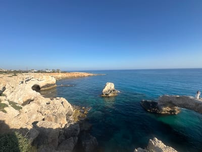 The image depicts a scenic coastal view in Ayia Napa, Cyprus. In the foreground, rugged, sun-drenched rocks form the cliff face, with sparse vegetation growing between them. The turquoise waters of the Mediterranean Sea gently lap against the shore. Further out, the sea transitions to a deeper blue. A prominent rock arch, a natural bridge, spans the water on the right side of the image, with a person dressed in white standing on it, facing away from the camera and looking out at the sea. In the distance, along the coastline, there are buildings and palm trees, suggesting a developed area beyond the natural landscape. The sky is a clear, bright blue.