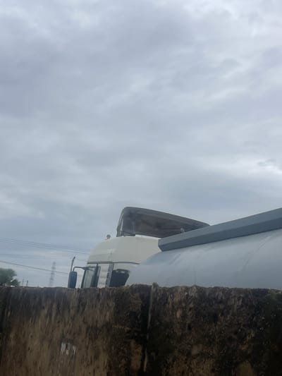 The image shows a view of the top portion of two tanker trucks partially obscured by a rough, textured concrete wall in the foreground. The sky above is overcast with thick, gray clouds. In the background, tall electrical pylons and some greenery are visible. The tanker trucks appear to be white with dark gray tops. One truck has a large, curved, silver tank, while the other has a white cabin with a dark gray cover over its top. The overall scene suggests an industrial or storage area in a cloudy environment.