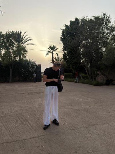 In Al Ouidane, Morocco, a person stands in a dusty courtyard during sunset. They are wearing a black t-shirt, white linen pants, and black shoes, with a black bag slung over their shoulder. The person is looking down at their hands, which are reaching towards their bag. In the background, a large metal gate is visible, with lush green bushes and trees on either side. Palm trees are scattered throughout the scene, and the sky is a hazy orange and yellow, indicating the sun is setting. Two other individuals are in the distance, one near the gate and another walking away from the camera. The ground is a textured, light brown dirt.