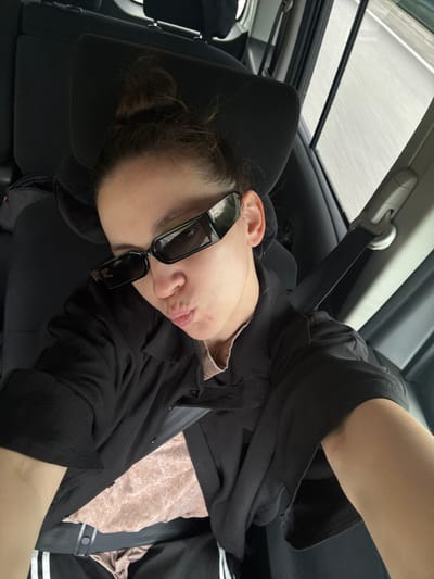A person is inside a car, leaning against the headrest. The person is wearing sunglasses and a black shirt, and a seatbelt is visible. The car has a sunroof. The location is not possible to determine.