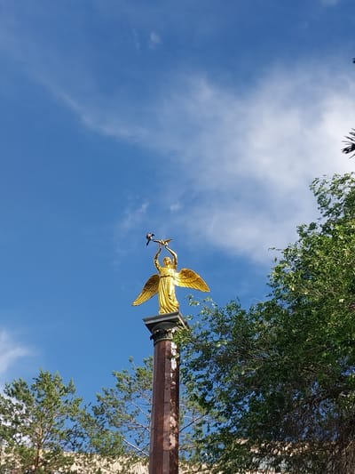 A golden statue of an angel stands atop a marble column, holding two birds in its outstretched hands. The statue is a prominent feature of the city of Staritsa, Russia. The angel is depicted with large, feathered wings and is wearing a flowing gown. The column is adorned with intricate carvings and appears to be made of polished marble. The background features a clear blue sky with a few wispy clouds, and lush green trees flank the statue. The overall scene is serene and picturesque, suggesting a monument of some significance within the city.