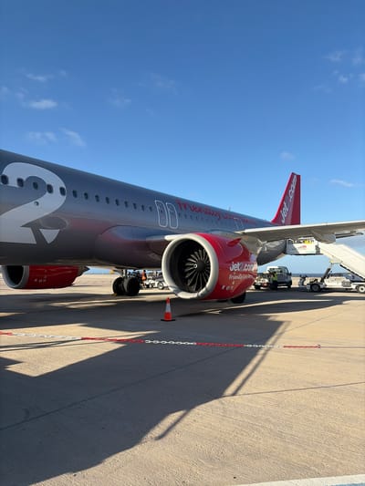 The image depicts a Jet2.com airplane parked on a concrete apron, presumably at an airport. It's a clear, sunny day with a blue sky. The plane is oriented lengthwise in the frame. A jet bridge is connected to the aircraft. Ground service vehicles and personnel appear to be assisting with boarding or other operations. A safety chain with white and red sections runs parallel to the aircraft. The shadow of the aircraft is cast on the apron. The engine reads "Friendly low fares". While there's no explicit visual confirmation of Puerto del Rosario, Spain, this is an assumption based on the provided context.