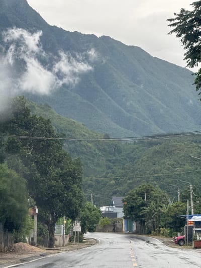 The image shows a landscape near Bình Lư, Vietnam, with a prominent mountain range partially obscured by low-lying clouds. A wet, paved road runs along the right edge of the frame, with intermittent yellow markings and a concrete barrier. On the roadside, there are utility poles with wires, buildings, vegetation, and a red vehicle.