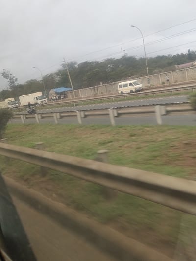The image depicts a multi-lane highway in Kahawa Wendani ward, Kenya, under an overcast sky. In the immediate foreground, a metal guardrail exhibits motion blur, indicating the photograph was taken from a moving vehicle. Beyond this, a grassy median separates the foreground from the main highway lanes. On the far left side of the road, several white box trucks are parked or moving slowly near a blue tent structure. A group of approximately 10-15 individuals is gathered around or under this blue tent. A motorcyclist is visible on a lane adjacent to the inner guardrail. Further down the highway on the right, a light-colored minivan is in motion. Two pedestrians are walking on a dirt path alongside a concrete barrier wall that separates the road from a treeline and what appears to be distant structures. Street light poles are spaced along the highway.