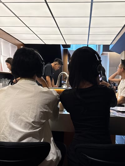 In the city of Jing'an District, China, two individuals are seated at a counter, both wearing headphones and facing away from the camera. The person on the left wears a white, button-down shirt, while the person on the right wears a black t-shirt. Behind them, another person in a black t-shirt stands and appears to be looking down at something on the counter, near a sink faucet. Further in the background, a person wearing glasses is visible on the right side of the frame. The environment appears to be a brightly lit indoor space with a modern ceiling design.